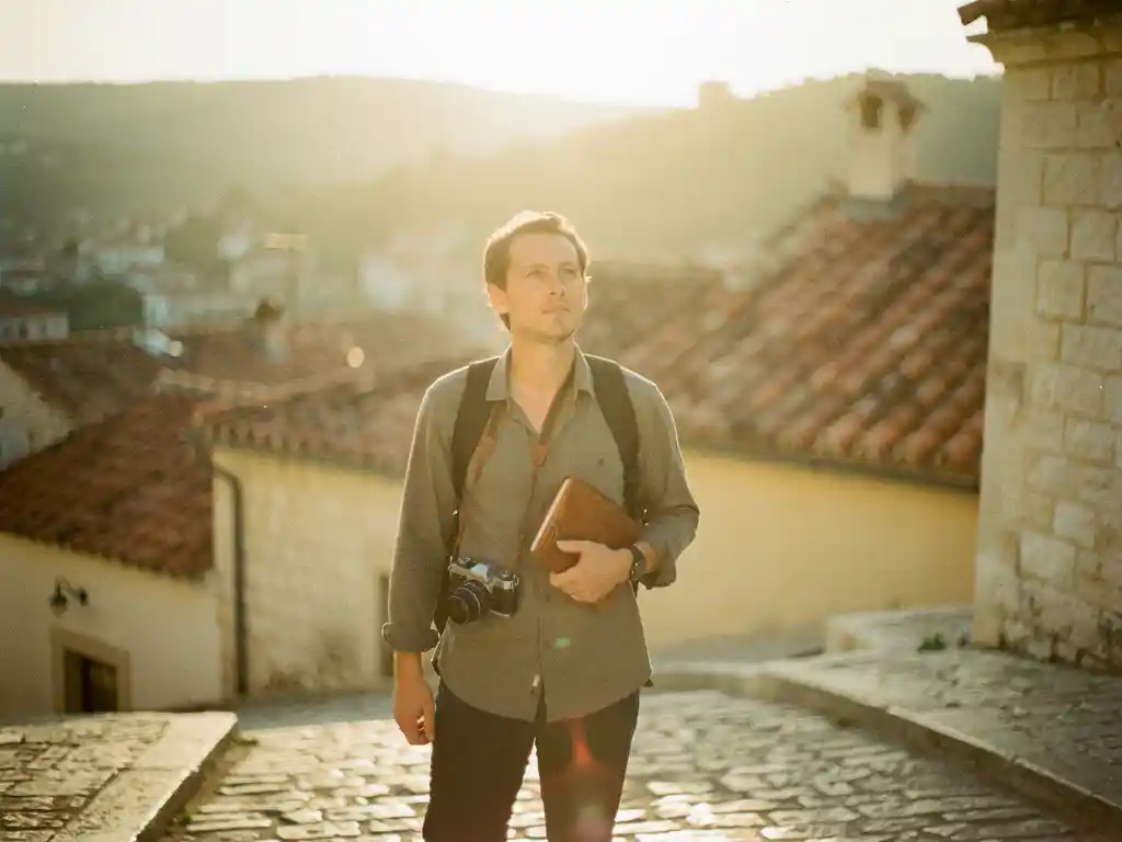 Traveler on sunlit Mediterranean cobblestone street holding vintage camera, terracotta rooftops and golden-hour hills in warm amber haze.